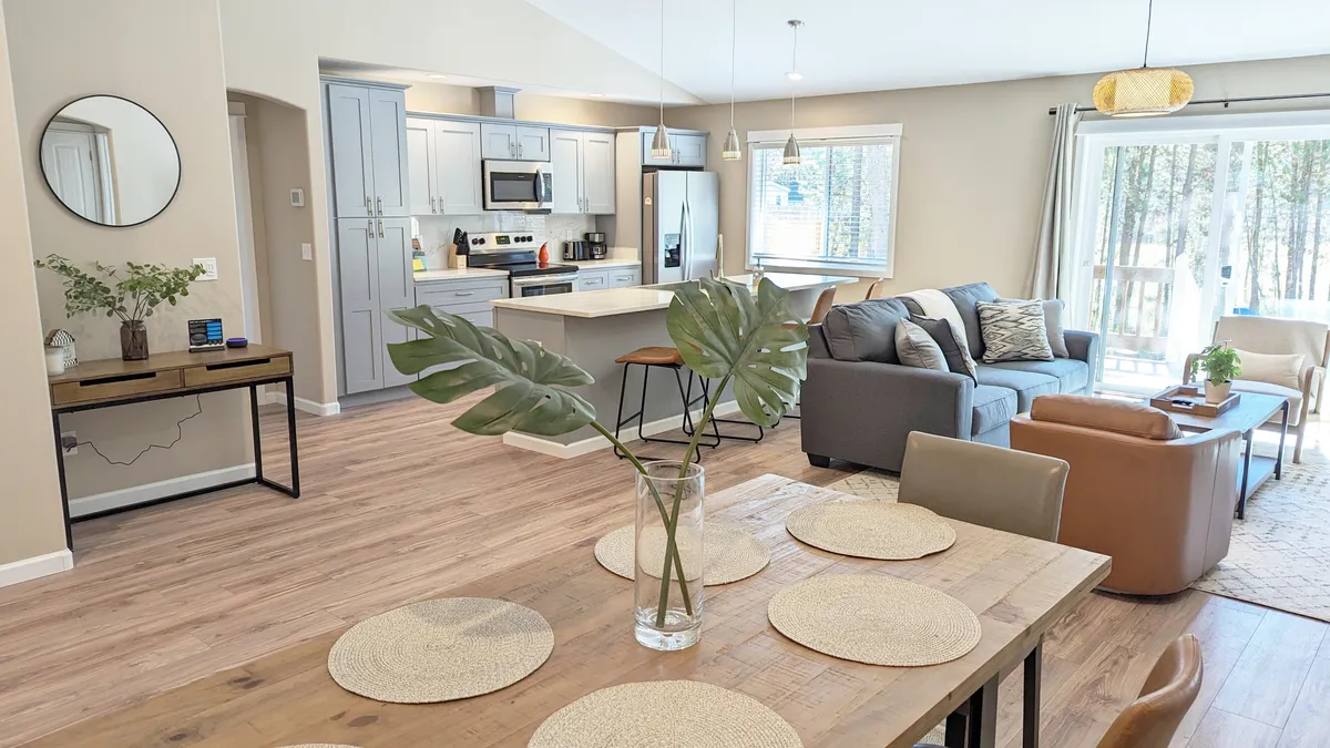 A cozy, open-concept living and dining area with a dining table set with placemats, a plush sofa seating area, and kitchen appliances visible in the background. Large windows suggest ample natural light and views of the surrounding pines, reinforcing the home’s connection to nature.