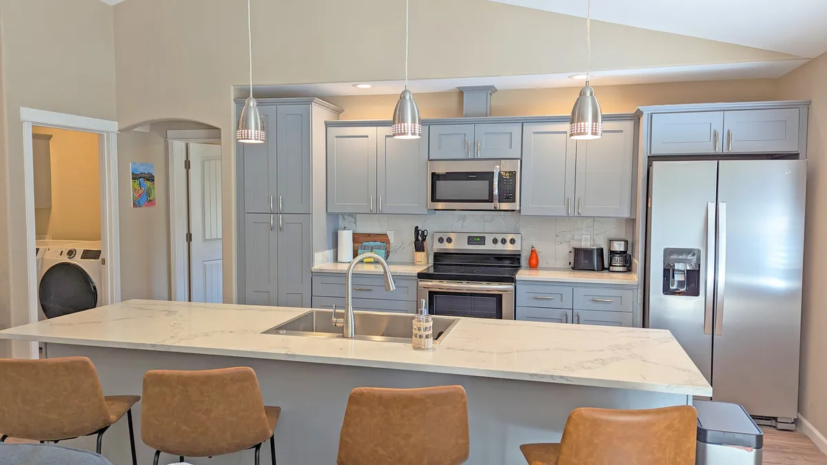 A stylish kitchen scene featuring a large quartz-veined island, stainless steel appliances, pendant lighting, and modern furnishings. The kitchen is well-lit and designed for both socializing and cooking, with bar stools inviting guests or family to linger and chat.