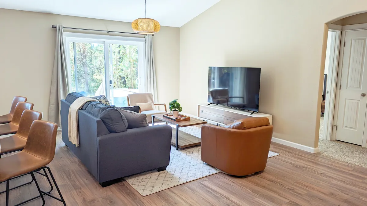The image showcases a bright, welcoming living room featuring a plush sofa, stylish coffee table, large windows that let in natural light, and a flat-screen television. The ambiance is cozy and perfect for unwinding or gathering with friends and family. The scene exudes comfort and relaxation, ideal for socializing, enjoying a movie night, or savoring quiet moments with a book and morning coffee.