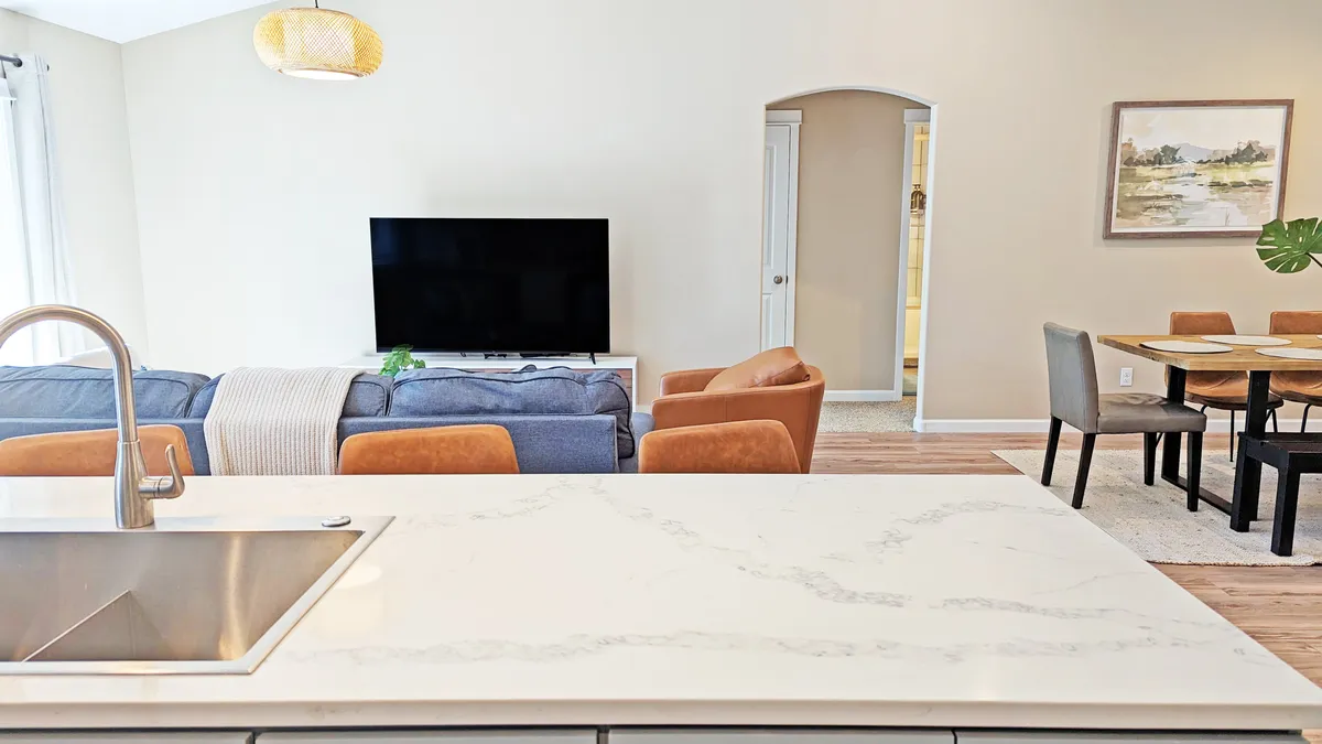 This image captures an inviting open-concept space featuring a modern kitchen island, a cozy sofa area, a large television, a dining table, and wall artwork. The space is bright and stylish, emphasizing togetherness and comfort. The kitchen is equipped with stainless steel appliances and ample seating at the island, while the living area offers a plush spot for relaxing or watching TV.