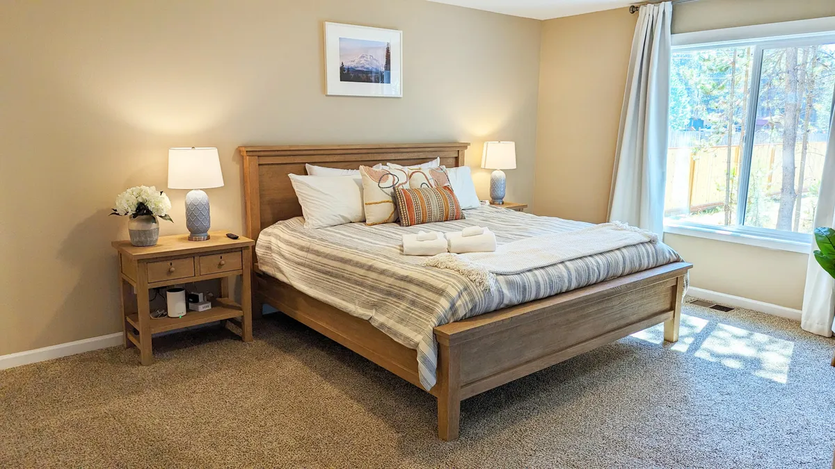 A tranquil bedroom featuring a neatly made bed, nightstand with a lamp, a window allowing natural light to pour in, and decorative wall art that adds a touch of personality. The soft, inviting atmosphere hints at comfort and privacy—ideal for a restful night's sleep.