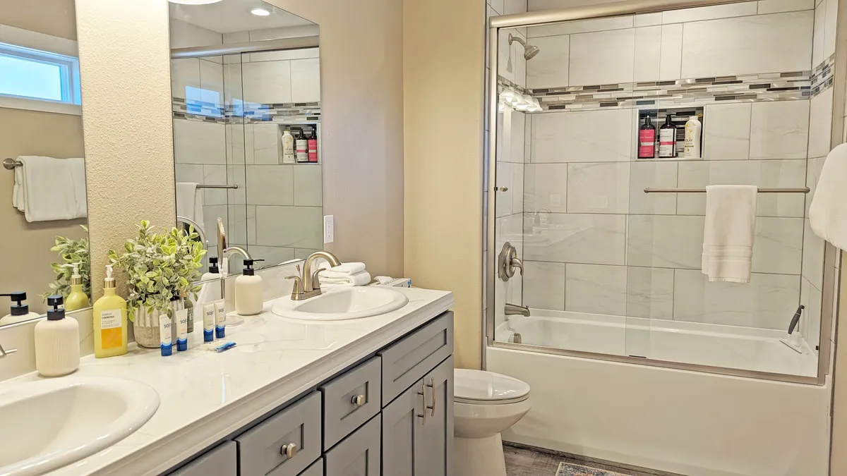 A spacious bathroom with a modern double sink vanity, framed mirror, walk-in shower, and elegant decorative touches. The space feels spa-like and luxurious, ideal for unwinding after a long day.