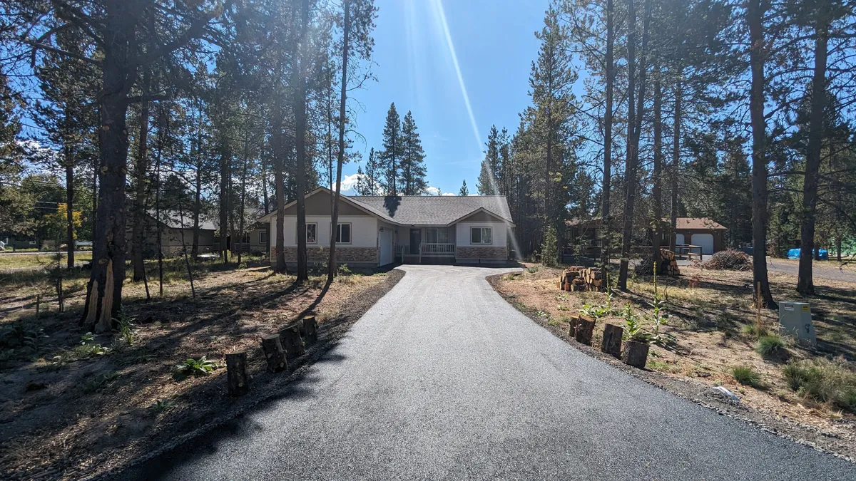 An exterior view of the house, featuring the driveway and surrounding trees, encapsulating the peaceful, wooded neighborhood setting. The scene evokes serenity and privacy, with the home nestled among mature pines.