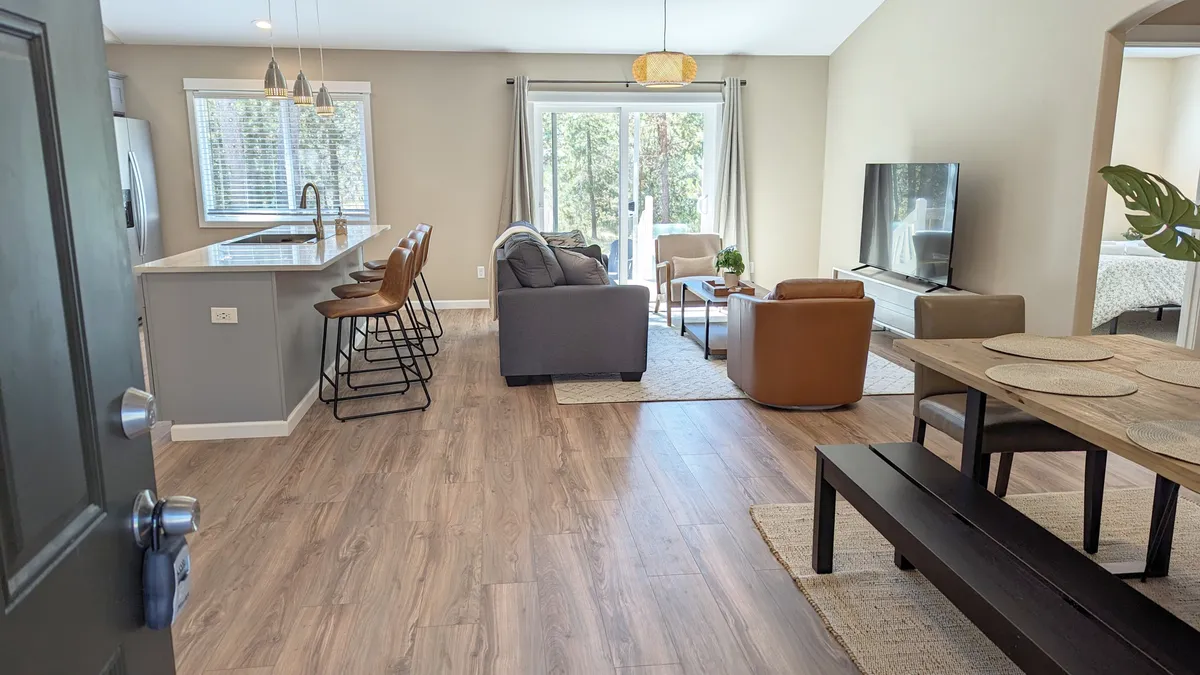 The open living area boasts an island bar with stools, a cozy sofa seating area, a dining table, and large glass doors leading to the outdoor space. The room is sunlit and spacious, ideal for lively gatherings, drinks, and group relaxation after outdoor adventures.