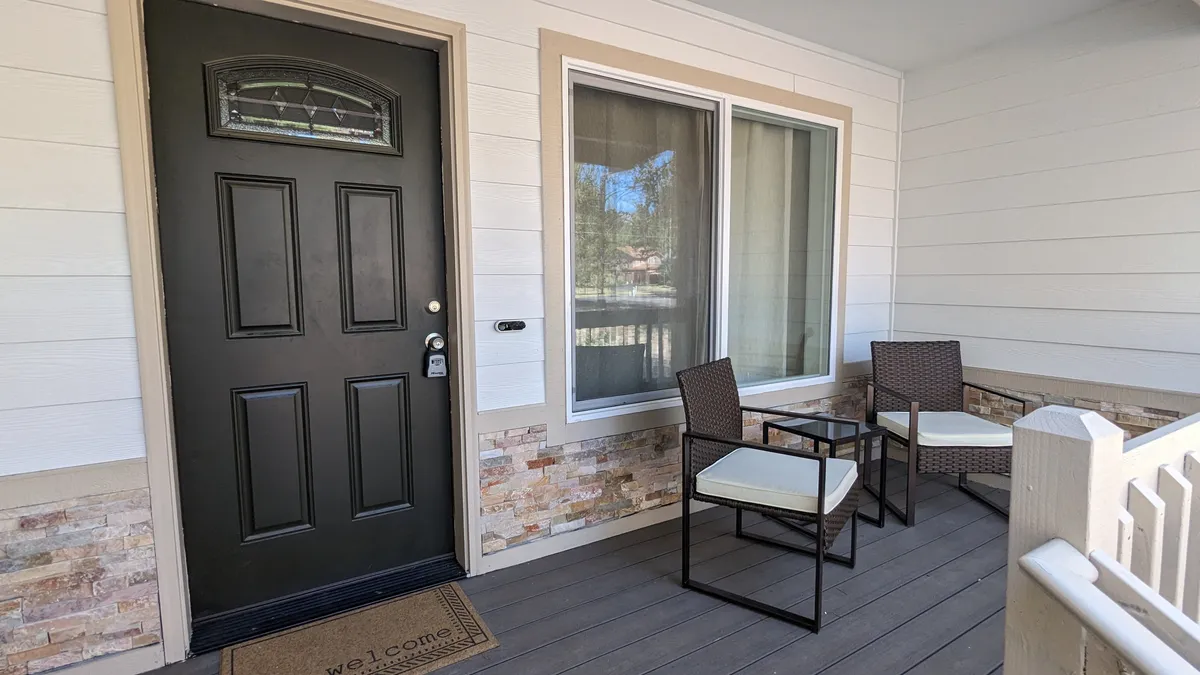 This exterior image showcases the home's welcoming entrance, featuring a black front door, a welcome mat, and a cozy seating area. The setting hints at a social space where friends greet each other as they arrive for a getaway, and the inviting atmosphere connects the indoors with the outdoors.