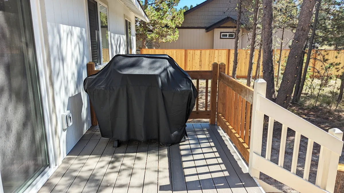 A propane grill (covered) is situated on a wooden deck with railings, surrounded by tall trees. The setting suggests outdoor meals and gatherings in a serene, natural environment.