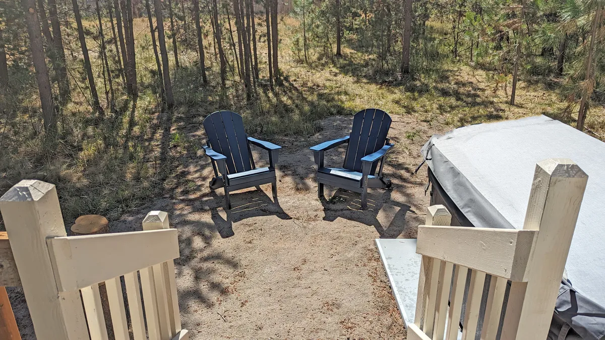 Two blue Adirondack chairs sit on a pathway surrounded by natural landscaping and tall pines, offering a peaceful spot to relax or socialize outdoors. The area feels private and immersed in nature—perfect for quiet moments or planning the next family outing.