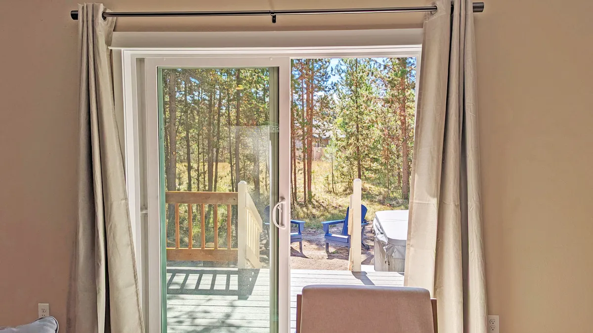 A view from inside the home looking out through a sliding glass door to a wooden deck with outdoor seating, nestled among trees. The deck offers a seamless connection between indoor comfort and outdoor fun, hinting at BBQs, wildlife watching, or family gatherings.